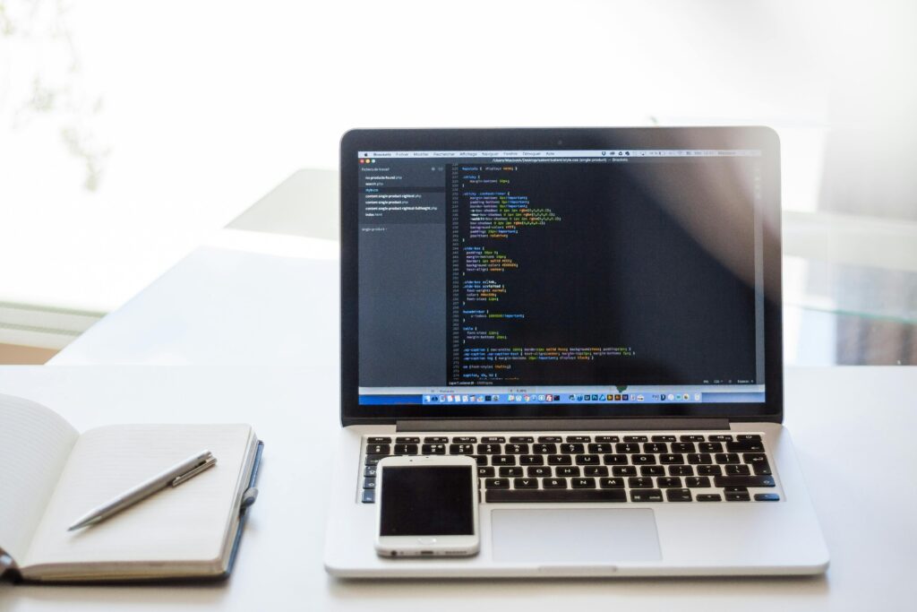 Open laptop displaying code on a desk with smartphone and notebook, ideal for technology and business use.